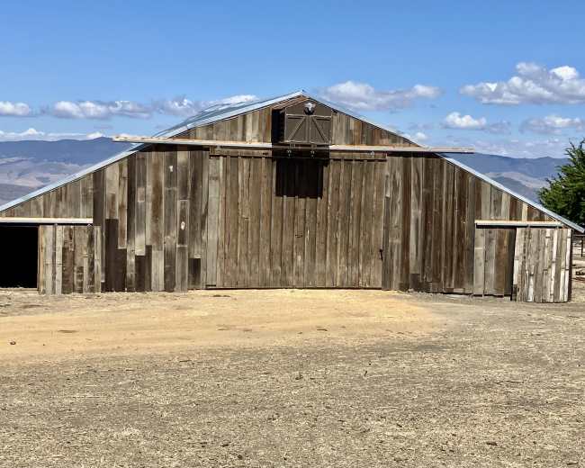 A large wooden barn with a sloped roof sits on a dirt lot, surrounded by hills and blue skies.