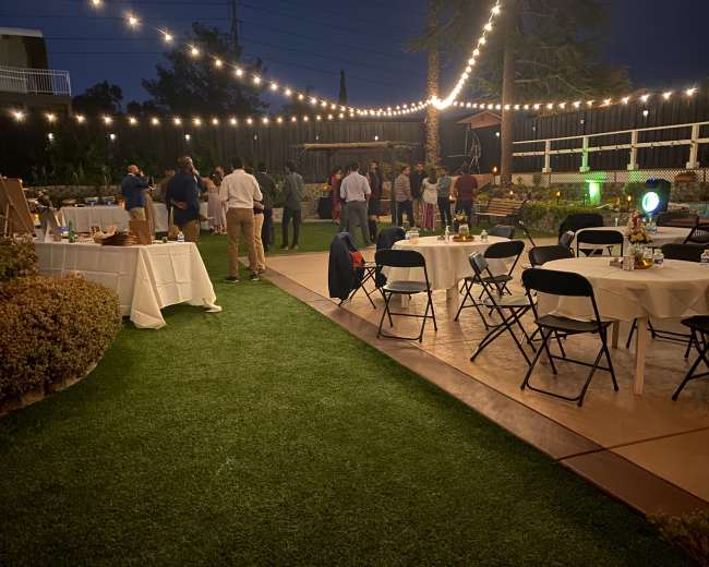 The scene shows a backyard party at night with string lights illuminating guests mingling near tables set up around a green lawn.