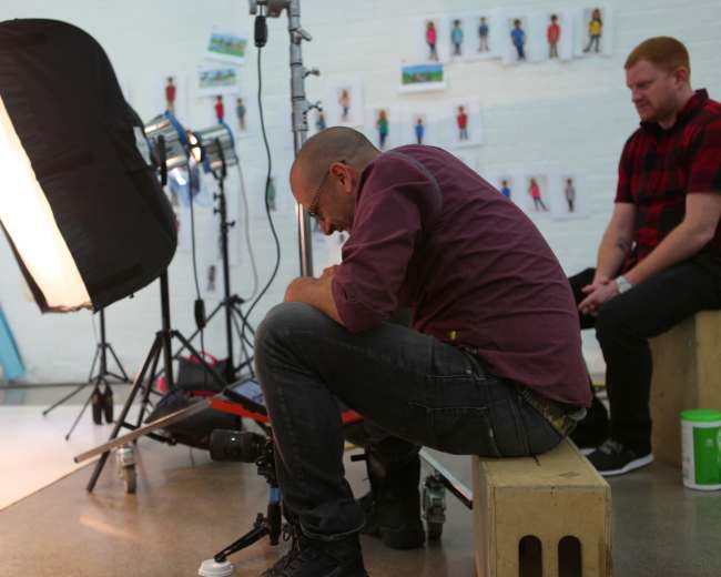 A man sits on a wooden bench, looking down in a studio filled with photography equipment and backdrops featuring model images on the wall.