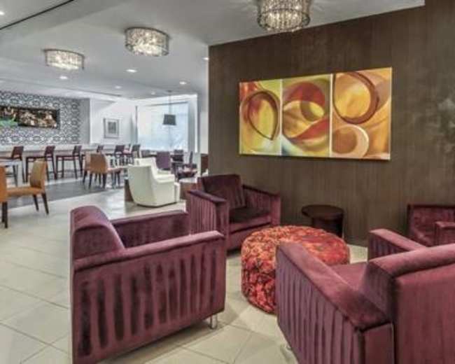 The image depicts a modern lounge area featuring sleek purple armchairs, a round patterned ottoman, and decorative wall art under stylish light fixtures.