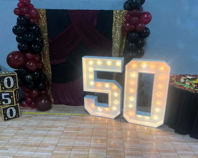 The image shows a decorated event space featuring large illuminated numbers "50," a backdrop with draped fabric and balloons, and a table with a floral arrangement.