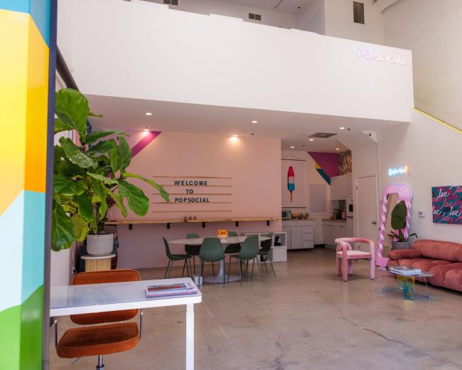 The image shows a colorful interior of a modern social space featuring a welcome sign, seating areas, and a vibrant decor.