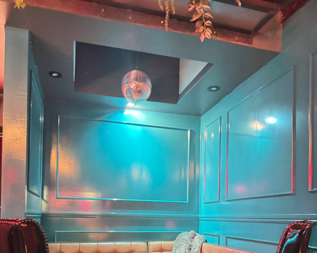 A cozy corner seating area features a beige padded bench and two black high-backed chairs, illuminated by a soft candlelight and colored lighting.