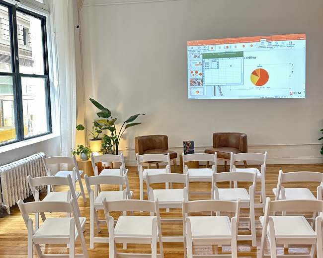 A room with white chairs arranged in rows facing a projector screen displaying a chart, with two brown chairs and a potted plant in the background.