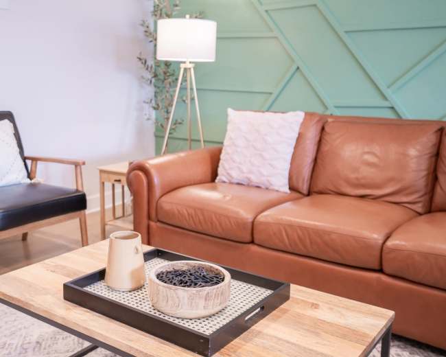 The image shows a living room setting with a brown leather sofa, a black armchair, and a wooden coffee table topped with decorative items.