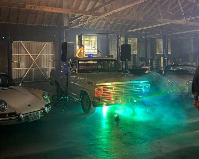 A vintage pickup truck with colorful lights is parked inside a warehouse filled with classic cars, while fog and ambient lighting illuminate the scene.