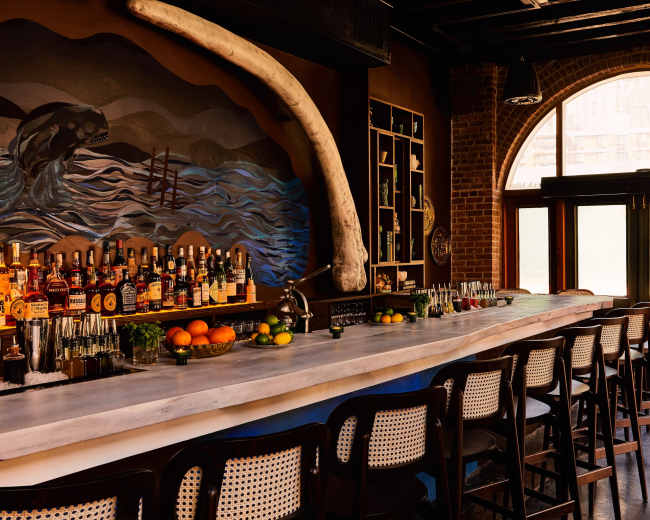 The image shows a stylish bar with a marble countertop, a selection of bottles on display, and large windows allowing natural light to fill the space.