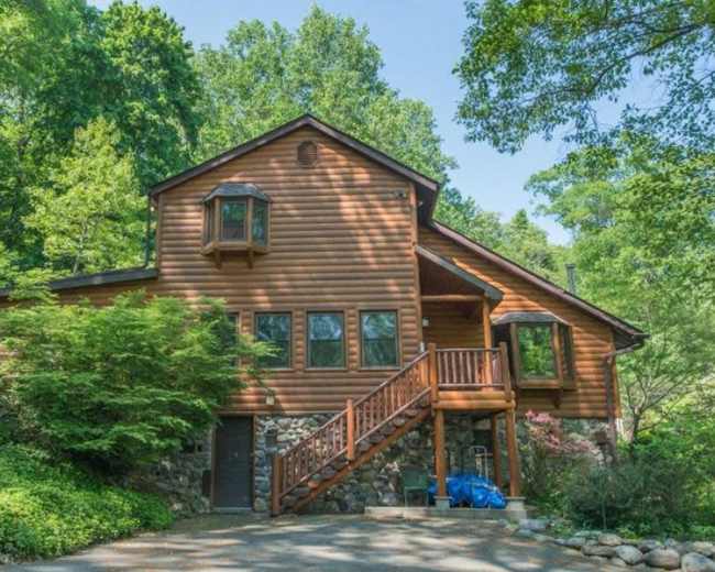A wooden cabin with large windows and a stone foundation is set amidst lush greenery.