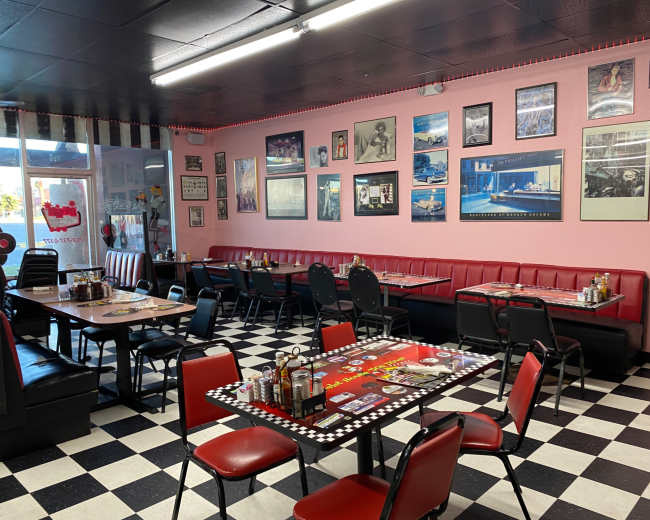 The image shows a retro-style diner with checkered black and white flooring, red booths, and tables adorned with condiments, featuring walls decorated with framed photographs and memorabilia.