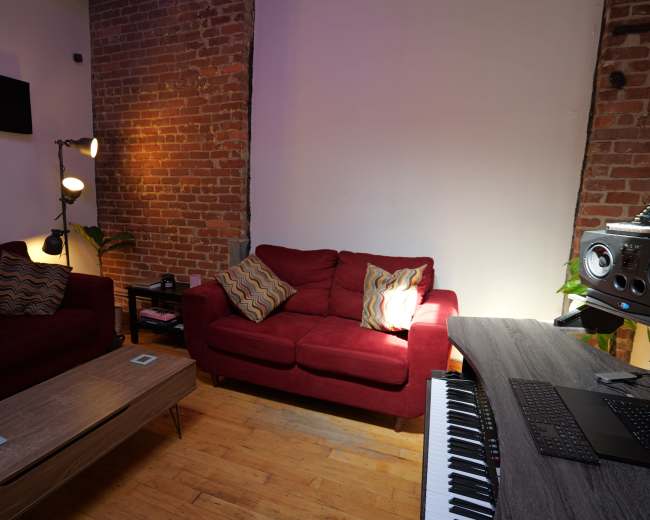 The scene features a cozy living room with red sofas, a wooden coffee table, and musical equipment on a desk against a backdrop of exposed brick walls.