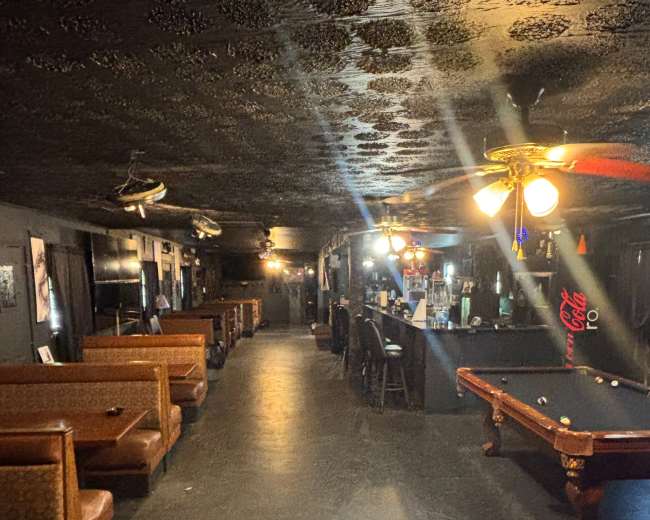 The image shows a dimly lit bar interior with seating booths along one side and a pool table near the center.