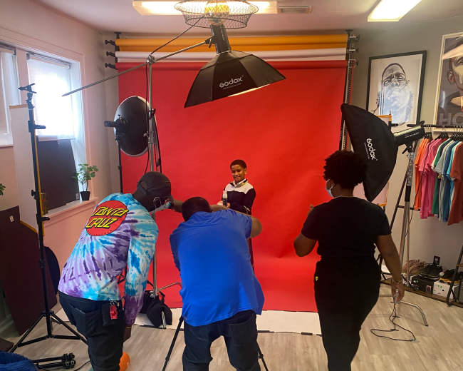 A child poses for a photoshoot in front of a red backdrop while three adults, equipped with cameras and lighting, adjust the setup.