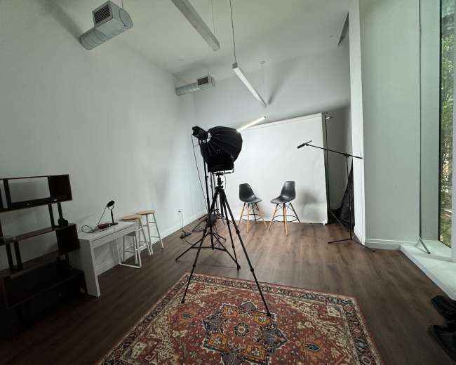 The image shows a bright, empty studio space featuring two chairs, a backdrop, and lighting equipment, with a wooden floor and a large window.