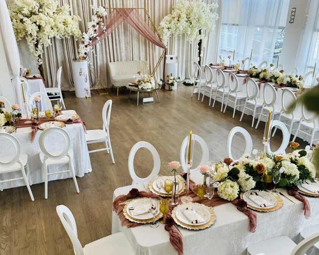 A elegantly arranged banquet hall featuring tables with white tablecloths, floral centerpieces, and decorative elements set for an event, with additional seating around the room.