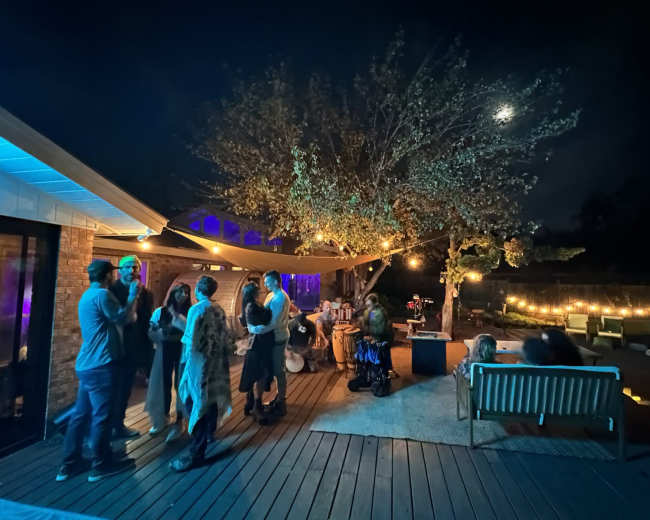 A group of people socialize on a wooden deck illuminated by string lights under a night sky with a full moon.