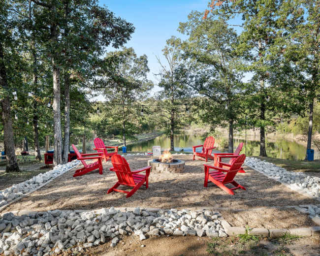 A circular seating arrangement of red Adirondack chairs surrounds a fire pit on a gravel and stone-lined area, set in a wooded location near a calm body of water.