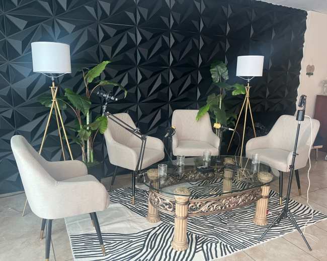 The image shows a seating area with four beige chairs arranged around a glass coffee table, set against a black 3D-patterned wall, with two floor lamps and a plant.
