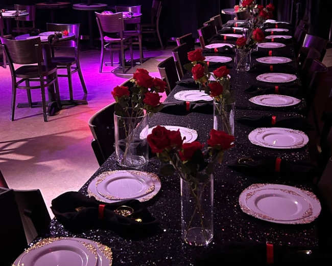 A long dining table is elegantly set with white plates, black napkins, and vases of red roses, beneath a dimly lit atmosphere adorned with decorative lighting.