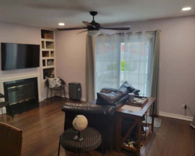 The image shows a living room with a dark leather couch, a wooden side table, a television mounted on the wall, and a window with sheer curtains.