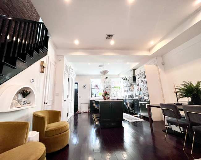 The image shows a modern kitchen and dining area with a staircase, featuring dark wood flooring, green cabinets, and a dining table with gray chairs.