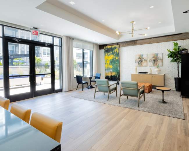 A modern lounge area with a seating arrangement, a small table, and a decorative wall art piece, all set against large windows and a light-colored brick wall.