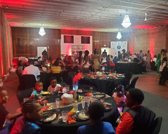 A large room is set up for a celebration, featuring tables with black tablecloths, decorated with centerpieces, while groups of guests socialize and enjoy food.