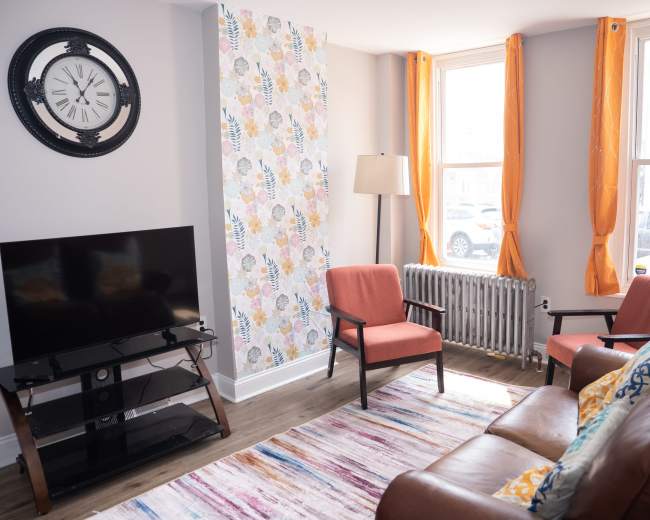 A living room features a television, two armchairs, a brown sofa, a patterned accent wall, and large windows with orange curtains.