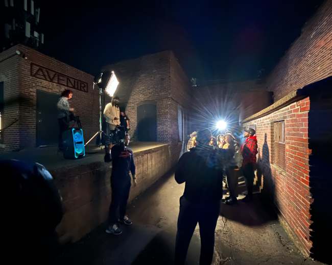 A group of people stands in a dimly lit outdoor area while a figure on a small platform speaks into a microphone, illuminated by bright lights.