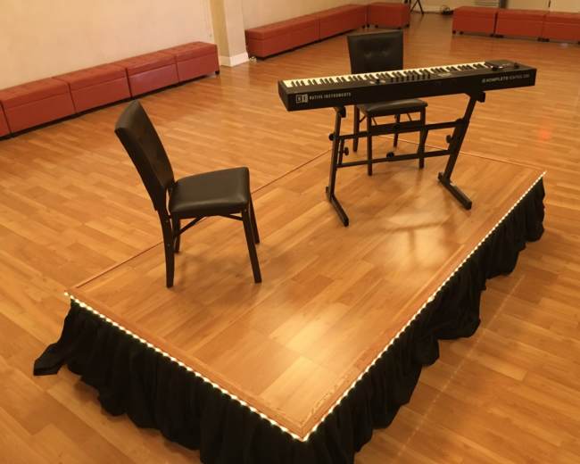 A keyboard and a chair are placed on a raised platform in a dance studio with wooden floors.