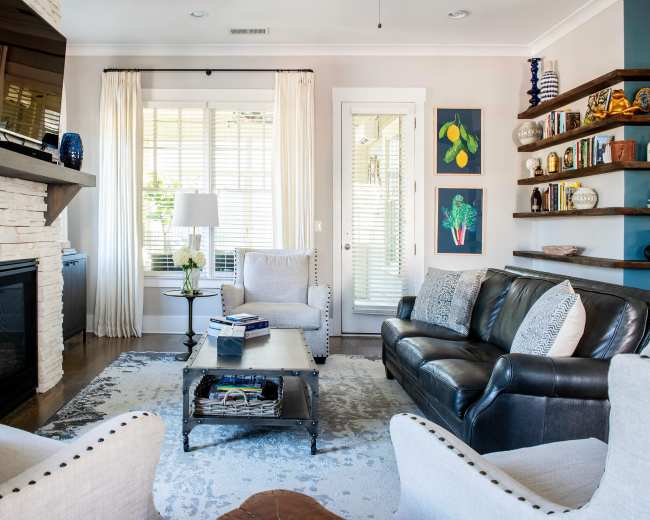 The image shows a modern living room featuring a fireplace with a flat-screen TV above it, two armchairs, a leather sofa, and a coffee table in the center, surrounded by decorated shelves and framed artwork on the walls.