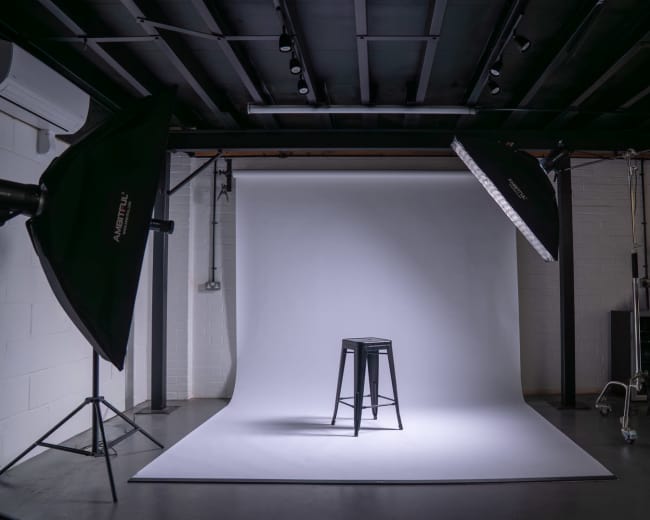 The image shows a photography studio equipped with two softboxes illuminating a black stool on a white backdrop.