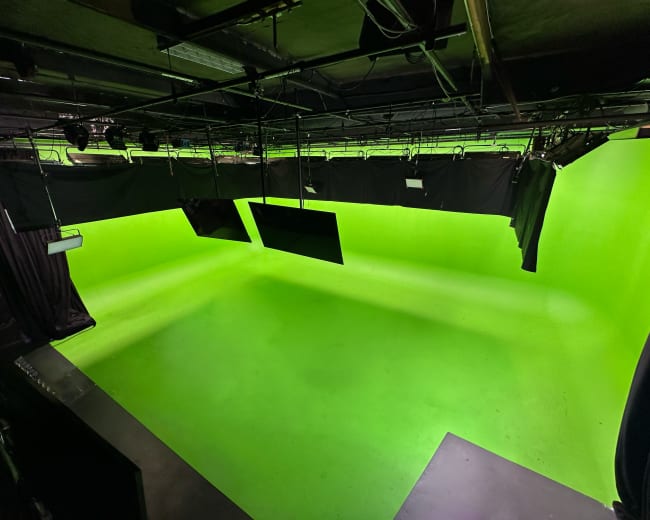 The image shows a green screen studio with a bright green backdrop and overhead lighting fixtures.