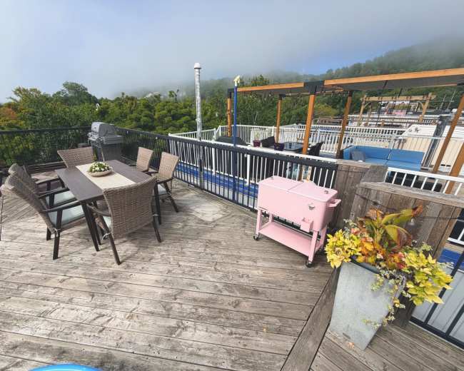 The image shows a rooftop patio featuring a wooden deck with a dining table and chairs, a pink cooler, and lush greenery in the background.