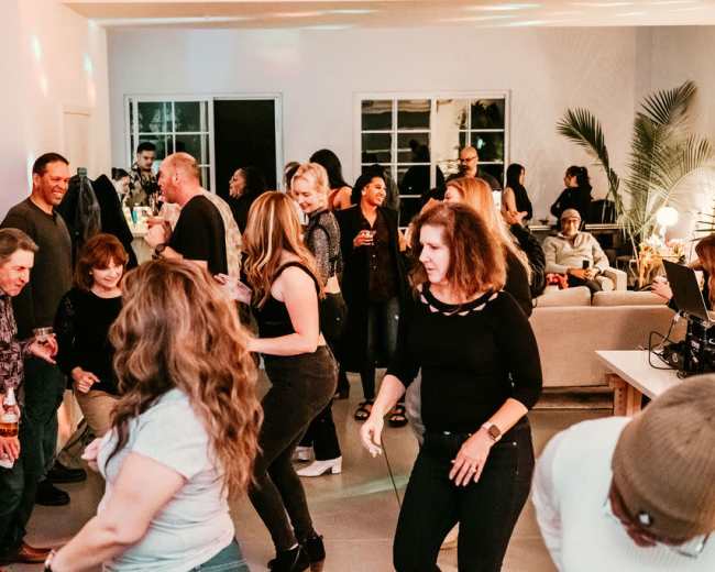 A group of people is dancing and socializing at a lively indoor gathering, with some seated in the background.