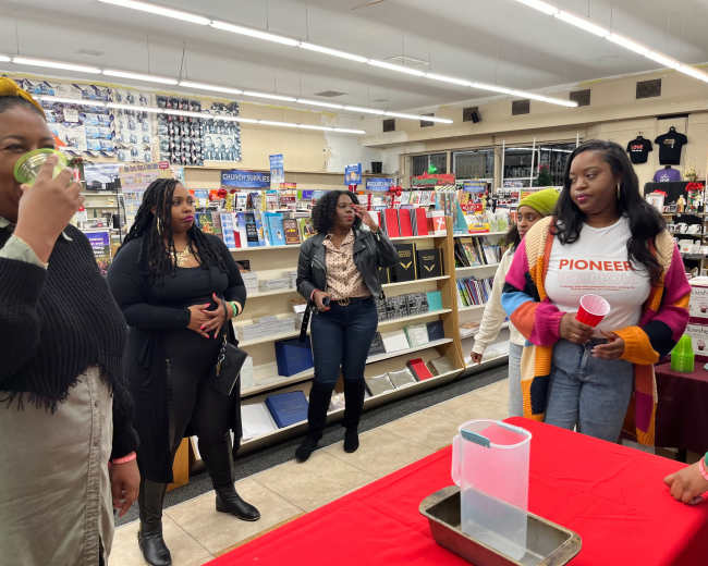 A group of people is gathered inside a store, interacting near a red table with a pitcher while shelves of various products are visible in the background.