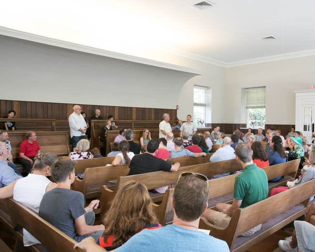 A crowd of people is seated in a wooden meeting hall while speakers stand at the front.