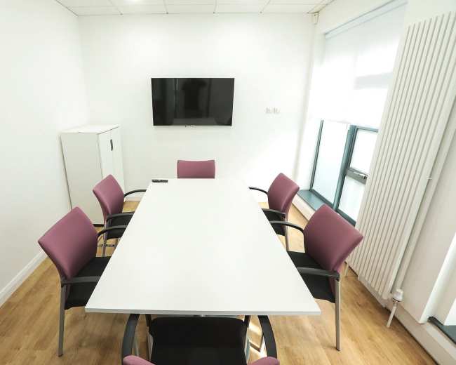 A conference room features a large white table surrounded by eight purple chairs and a wall-mounted television.