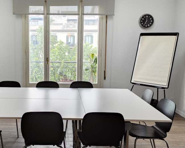 A bright meeting room features a large table surrounded by black chairs, with a clock on the wall and a flip chart nearby, overlooking a green view through large windows.