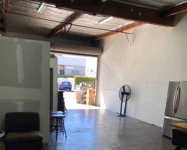 The image shows an empty industrial space with a concrete floor, a large wall fan, a kitchen area with a refrigerator, and a door leading to the outside.
