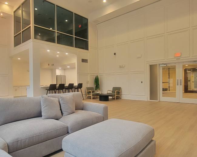 The image shows a spacious interior area featuring a light-colored sofa, a dining area with chairs, and a large windowed space upstairs.