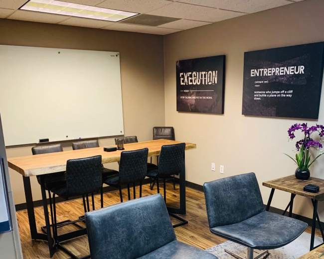 The image shows a modern conference room with a wooden table, black leather chairs, and motivational posters on the walls.