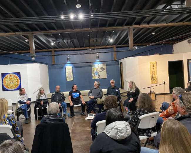 A group of eight people sits on chairs arranged in a semicircle in a spacious room with artwork on the walls, while an audience listens attentively.