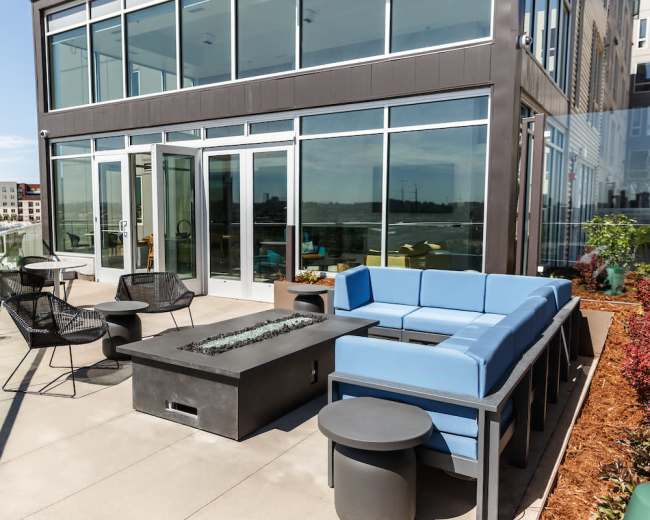The outdoor patio features a modern seating area with a blue sectional couch, a gas fire pit, and several black chairs and tables against a backdrop of large glass windows.
