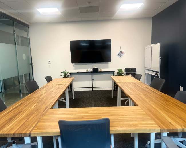The image shows a modern conference room with several wooden tables arranged in a U-shape around a large screen mounted on the wall.