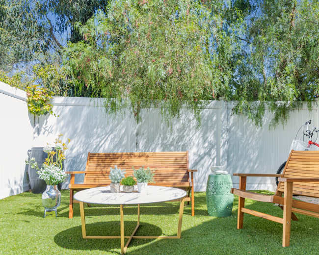 The image shows a sunny outdoor seating area with two wooden chairs, a round table, potted plants, and a bicycle leaning against a fence.