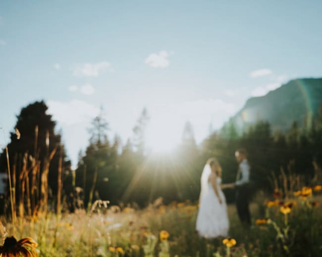 A bride and groom stand in a sunlit field surrounded by wildflowers and tall grass, with mountains in the background.