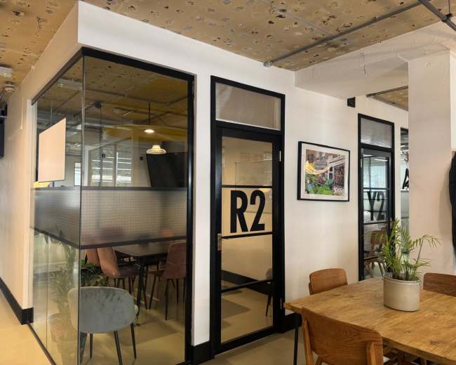 The image shows a modern office space with a glass-walled meeting room labeled "R2," surrounded by a dining area featuring wooden tables and various seating options.