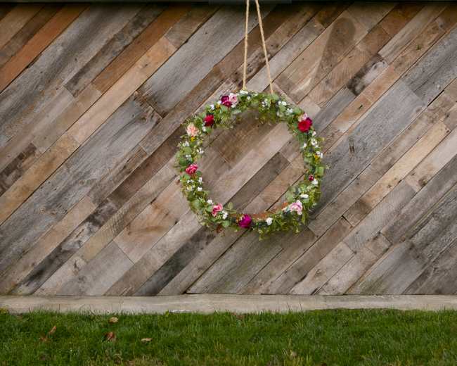 A circular flower wreath hangs on a textured wooden wall set against a grassy area.