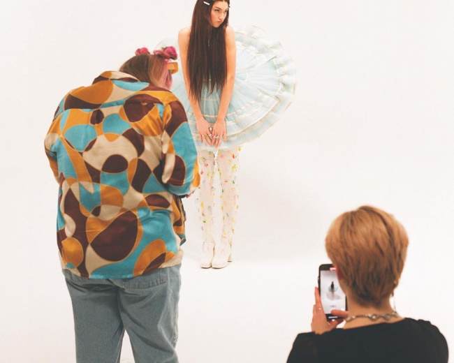 A model poses in a light blue outfit with a tulle skirt while two photographers capture the scene from different angles.