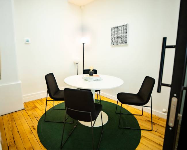A small meeting room features a round white table surrounded by four black chairs on a green circular rug, with a lamp and framed photograph on the walls.